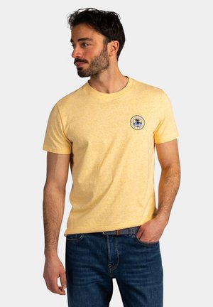 Man draagt een gele korte mouwen t-shirt met een klein rond palmboomlogo op de borst, gecombineerd met een blauwe spijkerbroek en een patroonriem.