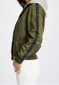 Olivgrön bomberjacka med grå huva, svart sidstripe med text, ribbade ärmslut och midja. Har dragkedjestängning och sidfickor.