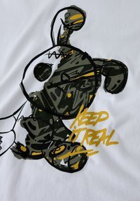 Boceto abstracto de un oso de peluche con patrones en estilo grafiti en negro y dorado y la frase "MANTENLO REAL" sobre un fondo de tela blanca.