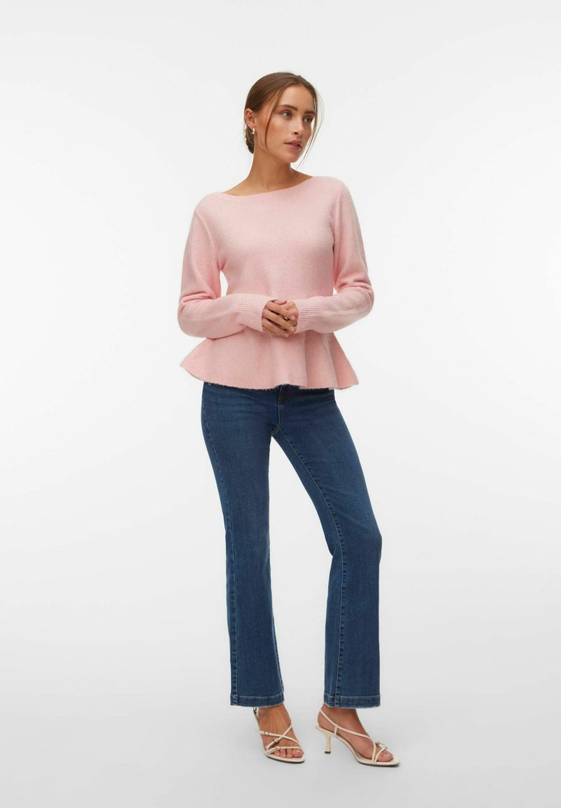 Femme portant un pull péplum rose à manches longues, un jean bleu bootcut et des sandales à talons beige avec des brides, debout les mains jointes.