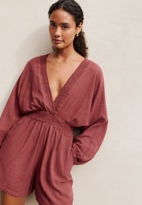 Maroon linen jumpsuit with deep V-neck, smocked waist, and loose long sleeves, featuring a textured fabric and relaxed fit.