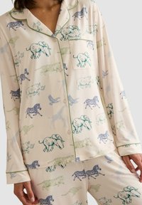 Conjunto de pijama en tejido crema con un patrón de varios animales, incluyendo elefantes, cebras y tigres, con ribete verde y un bolsillo de parche.