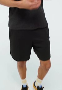 Pantaloni sportivi neri realizzati in tessuto leggero, con una cintura elastica. Abbinati a sneaker nere e calze bianche e gialle.