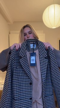 Houndstoothjaspatroon in zwart en crème, met een reverskraag, knopen en een beige voering. Prijsetiket bevestigd aan de binnenkant van de kraag.