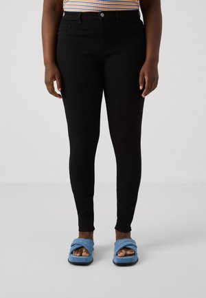 Persoon draagt zwarte skinny jeans gecombineerd met blauwe open-toe slides en een meerkleurige gestreepte top, staand op een lichte achtergrond.