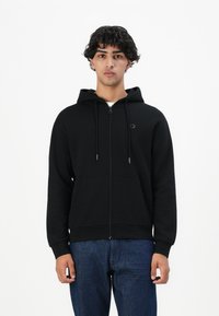 Ung mann med krøllete mørkt hår iført svart zip-up hettegenser og blå jeans, stående foran en hvit bakgrunn.