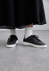 Zapatillas negras con patrón texturizado en relieve, diseño de cordones y suela de goma blanca, combinadas con calcetines blancos hasta el tobillo y una falda negra plisada.