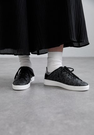 Zwarte sneakers met een textuur gegraveerd patroon, vetersluiting en een witte rubberen zool, gecombineerd met witte crew-sokken en een geplooide zwarte rok.