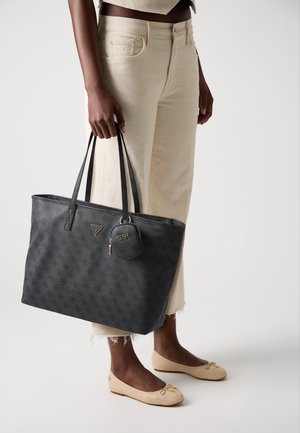 Grote zwarte tote bag met logopatroon, textuurmateriaal, dubbele handvaten en een afneembare kleine pouch. Vergezeld door beige flats en crèmekleurige broeken.