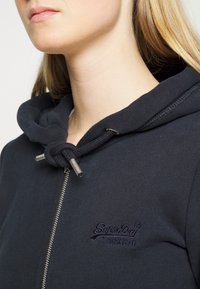 Marinefarvet zip hoodie med snoretræk i hætten, sølvfarvet lynlås og broderet "SuperDry" logo i mørk tråd på brystet. Glat stofstruktur.