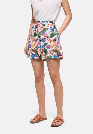 Mujer con shorts de talle alto con estampado tropical, camiseta blanca metida por dentro y sandalias marrones trenzadas, de pie con una mano en el bolsillo.