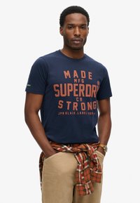 Camiseta de manga corta azul marino con el texto naranja "MADE MFG SUPERDRY CO STRONG." Combinada con pantalones beige y una camisa de cuadros atada alrededor de la cintura.