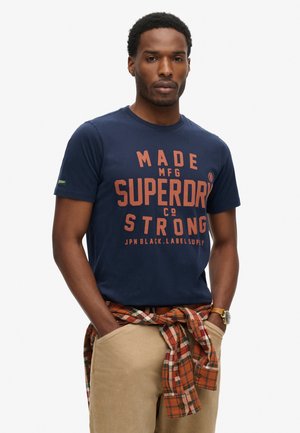 Granatowa koszulka z krótkim rękawem z pomarańczowym napisem "MADE MFG SUPERDRY CO STRONG." Połączona z beżowymi spodniami i w kratkę koszulą zawiązaną w talii.