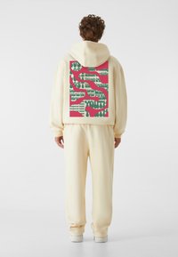 Cremefarvet hætte sweatshirt med et stort farverigt grafik på ryggen, der viser grøn tekst på en rød baggrund. Blødt stof og afslappet pasform.