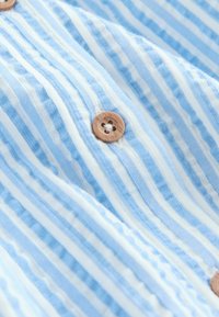 Hellblaue und weiße gestreifte Stoff mit einer strukturierten Haptik; verfügt über einen runden Holzknopf mit einem blumenförmigen Lochdetail.