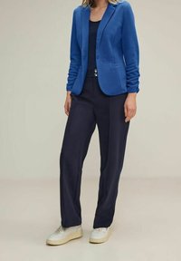 Blå kavaj i mjukt tyg, kombinerad med mörkblå byxor som har en avslappnad passform och slät textur. Vita sneakers fulländar outfiten.