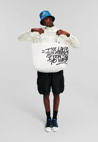 Borsa tote in tela con testo nero audace, indossata da una persona con una camicia bianca e pantaloni corti neri, accessoriata con un cappello a secchiello blu con fantasia e scarpe colorate.