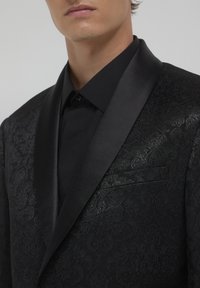 Giacca in tuxedo nera caratterizzata da un motivo damascato testurizzato, colletto revers in satin e taschino a petto singolo, abbinata a una camicia nera semplice.