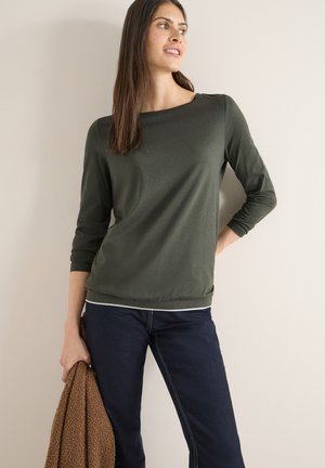 Femme en haut vert à manches longues et jean foncé tenant une veste marron texturée, debout contre un mur clair uni, regardant sur le côté.