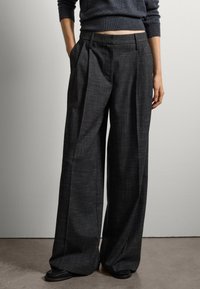 Brede, antracietgrijze broek met voorplooien, textuurstof en zijzakken. Geddragen met een donkere trui en platte schoenen.