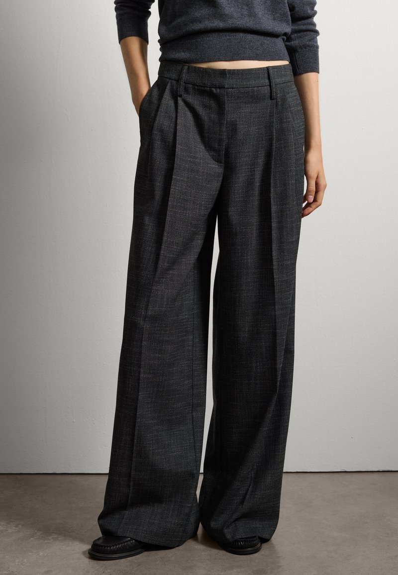 Brede, antracietgrijze broek met voorplooien, textuurstof en zijzakken. Geddragen met een donkere trui en platte schoenen.