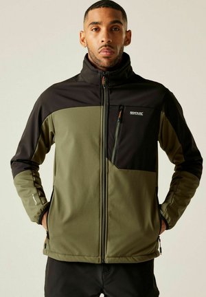 Hombre con chaqueta de exterior con cremallera en verde oliva y negro, con las manos en los bolsillos, mirando al frente sobre un fondo liso.