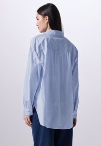 Camicia a maniche lunghe azzurro chiaro con yoke posteriore, polsini abbottonati e sottili righe verticali. Vestibilità comoda con orlo curvo.