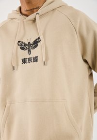 Beige hoodie med sort møl grafik og japansk tekst, båret af en person med en "tro på dig selv" hals-tattoo.