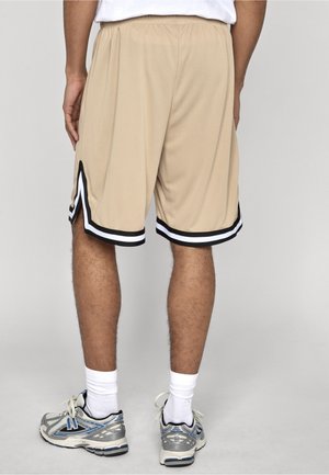 Man wearing beige basketball shorts with black and white trim, white socks, and silver athletic shoes standing against a neutral background.