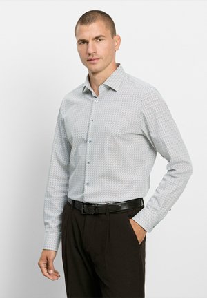 Man met kort haar die een lang mouwig overhemd met patroon en donkere broek met een zwarte riem draagt, staand voor een effen achtergrond.