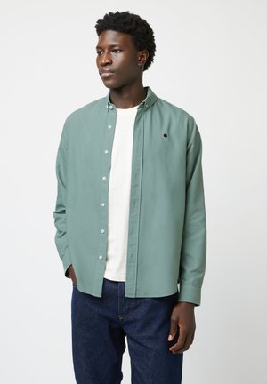 Jeune homme portant une chemise boutonnée vert clair sur un T-shirt blanc et un jean bleu foncé, debout devant un fond uni clair.