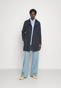 Tumed kahepoolselt kinnitatav trench coat suurte nuppudega; kantud helesinise särgiga ja laia säärega teksapükstega; minimalistlik disain ja tekstuuriga kangas.