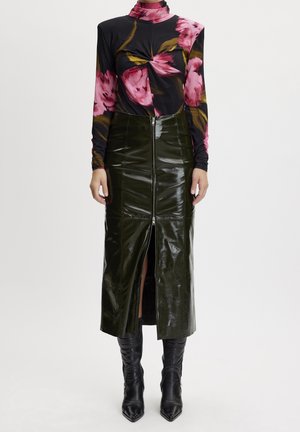 Mujer con un suéter de cuello alto negro con estampado floral rosa, una falda midi de cremallera al frente de color verde oscuro brillante y botas negras de punta fina.