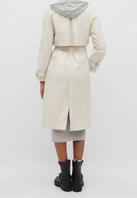 Osoba ubrana w beżowy płaszcz trenchowy z paskiem i rozcięciem z tyłu, szarą sukienkę z kapturem oraz czarne botki za kostkę, stojąca na jednolitym tle.