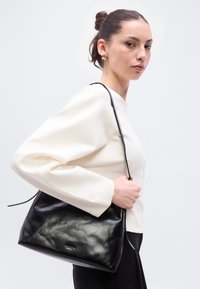 Μαύρη δερμάτινη τσάντα χειρός με απαλό, υφή επένδυση, τριγωνικό σχήμα και μακρύ λουρί, κρατημένη από ένα άτομο που φοράει λευκό τοπ.