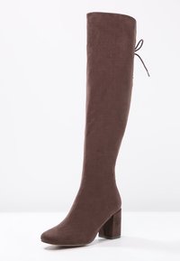 Bota marrón por encima de la rodilla hecha de suave ante, con un tacón cuadrado y detalle de lazo ajustable en la parte superior. Textura suave, forma ajustada.