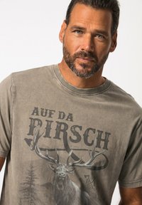 Camiseta gris de manga corta con un gráfico de un ciervo con grandes cuernos y el texto "AUF DA PIRSCH", hecha de una tela suave con una textura desgastada.