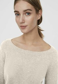 Pull en beige clair avec une texture douce et un décolleté bateau large, présentant une coupe légèrement ample et des bords côtelés.
