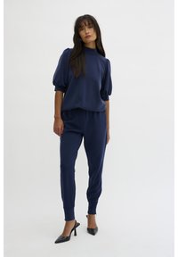 Navy blue loungewear set featuring a high neck top with puffed sleeves and tapered pants. Fabric appears soft and smooth. Worn with black heels.