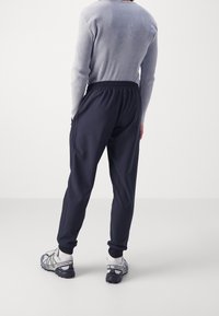 Osoba ve černých joggingových kalhotách, šedém žebrovaném dlouhém rukávu, bílých ponožkách a šedo-černých teniskách Salomon stojící na bílém pozadí.