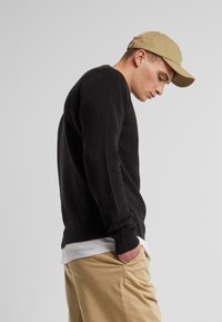 Black ribbed sweater with long sleeves, worn over a white shirt, paired with khaki pants and a beige cap. Simple, textured design.