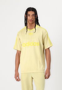 Sárga adidas póló függőleges csíkokkal, amelyen egy feltűnő "adidas" logó található. A vállakon kontrasztos három csík látható.