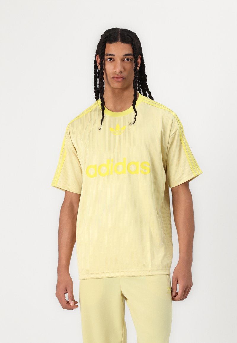 Sárga adidas póló függőleges csíkokkal, amelyen egy feltűnő "adidas" logó található. A vállakon kontrasztos három csík látható.