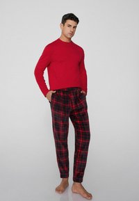 Červený top s dlouhým rukávem spárovaný s černými a červenými kostkovanými pyžamovými kalhotami s elastickým pasem. Model stojí bosý s rukama v kapsách.