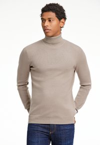 Hellbeige Rollkragenpullover mit geripptem Muster, langen Ärmeln und taillierter Passform. Kombiniert mit dunkelblauen Jeans.