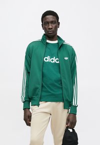 Grønn treningsjakke med hvite striper, glidelåslukking, ribbestrikkede mansjetter, lagd over en grønn t-skjorte med Adidas-logo, kombinert med beige bukser.