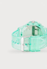 Transparent grön plastklocka med silverspänne, rund urtavla och synliga timmarkeringar. Har ett flexibelt armband med små nitar.