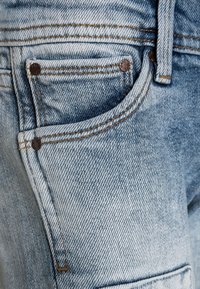 Vaqueros de mezclilla azul claro con un bolsillo frontal, remaches de cobre y costuras en contraste de color naranja, con una textura desvaída.