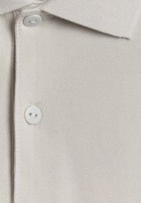 Tissu de polo beige avec deux boutons blancs et un col plié, montrant un matériau tricoté texturé.