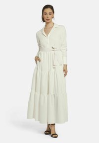 Langes weißes Kleid mit einem geknöpften Hemdkragen, Stufen am Saum, einem gebundenen Taillengürtel und seitlichen Taschen. Getragen mit schwarzen Absatzsandalen.